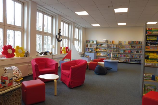 Innenansicht einer gemütlichen Leseecke in einer Bücherei mit roten Sesseln, bunten Kissen und Bücherregalen.