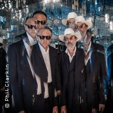 Mehrere Männer in Anzügen und Sonnenbrillen stehen vor einem verspiegelten Hintergrund.