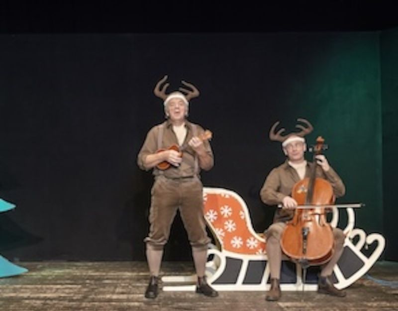 Zwei Männer in Rentierkostümen auf einer Bühne, einer spielt Ukulele, der andere Cello, neben einem Schlitten.