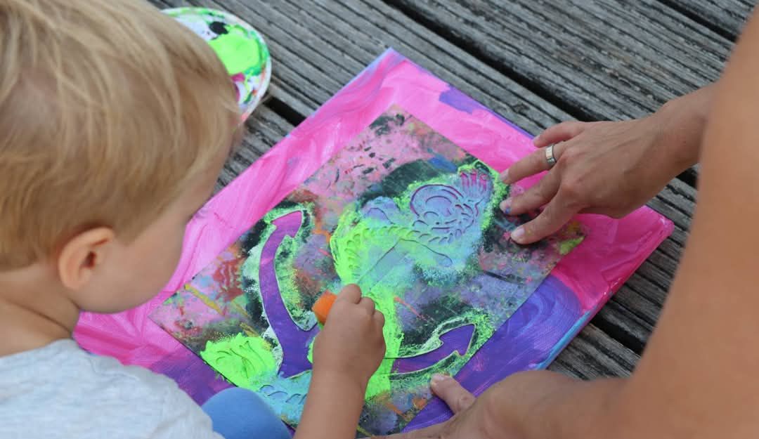 Ein Kind malt mit leuchtenden Farben auf einer Schablone, die auf einem Holzdeck liegt. Eine erwachsene Hand hilft dabei.