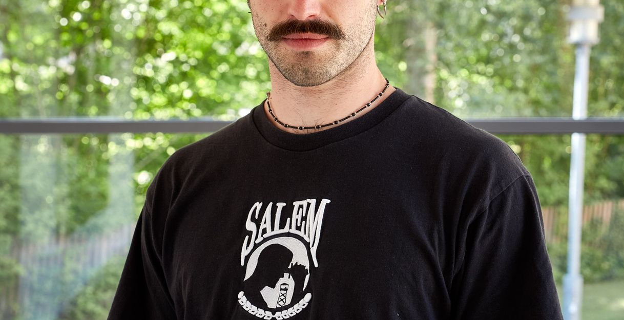 Einjunger Mann mit Schnauzer und Halbglatze in einem schwarzen Tshirt schaut direkt in die Kamera