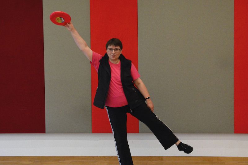 Eine Frau in Sportkleidung hält eine Frisbee und balanciert auf einem Bein in einem Raum mit bunten Wänden.