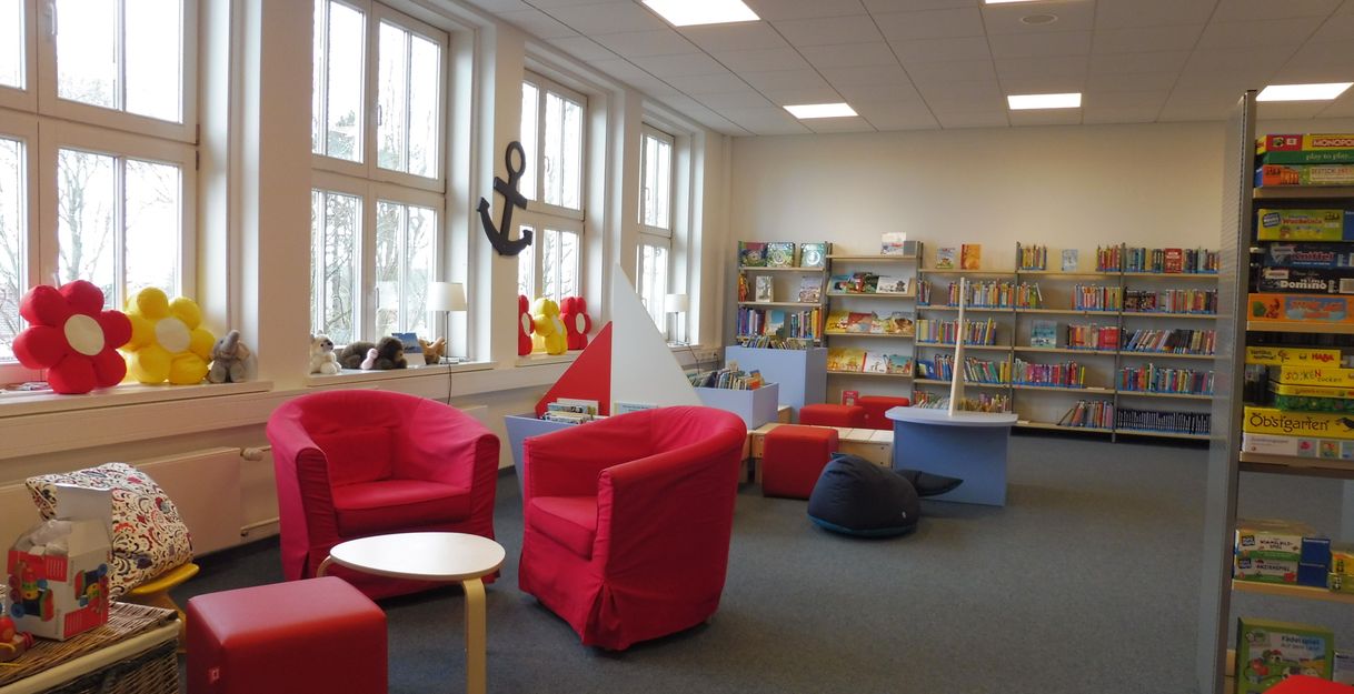 Innenansicht einer gemütlichen Leseecke in einer Bücherei mit roten Sesseln, bunten Kissen und Bücherregalen.