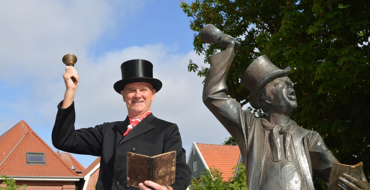 Ein Mann in schwarzem Anzug und Zylinder steht neben einer Statue, die ebenfalls einen Zylinder trägt. Beide halten eine Glocke und ein Buch in der Hand.