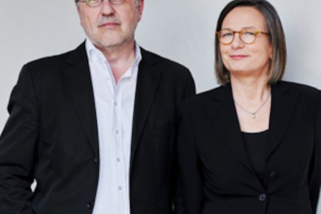 Ein Mann und eine Frau in formeller Kleidung stehen nebeneinander vor einem neutralen Hintergrund.