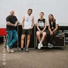 Vier Männer stehen und sitzen lässig vor einer weißen Wand, umgeben von Musikequipment.