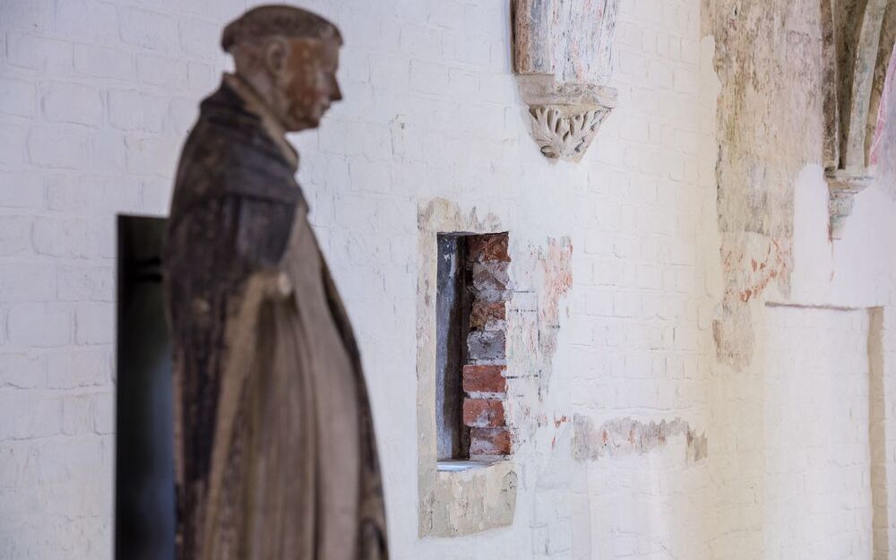 Foto in schräger Perspektive auf eine weiß gestrichene Backsteinwand., in die ein kleiner Durchbruch geschlagen wurde. Im Vordergrund steht eine Dominikanerstatue.