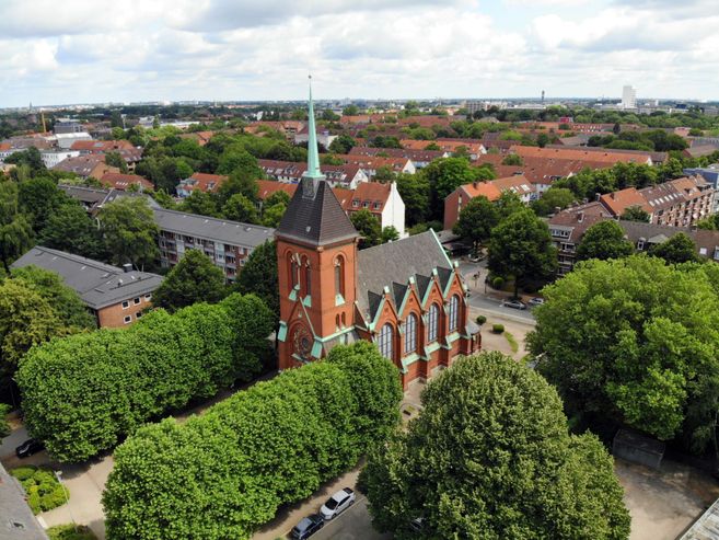 Luftaufnahme einer Kirche mit rotem Backstein und grünem Dach, umgeben von Bäumen und Wohnhäusern.
