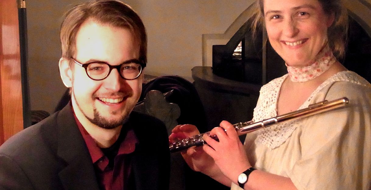 Ein Mann mit Brille und eine Frau mit Flöte lächeln in die Kamera.