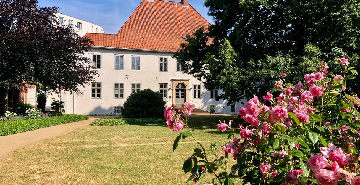 Historisches Gebäude mit rotem Dach und Garten mit blühenden Rosen im Vordergrund.