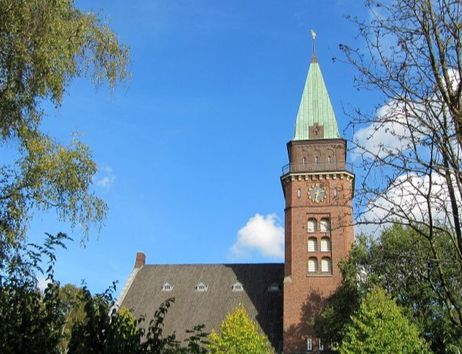 Ein Kirchturm mit grünem Dach ragt in den blauen Himmel, umgeben von Bäumen.