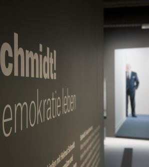Fotos, das eine von der Seite aufgenommene Informationstafel im Vordergrund zeigt. Darauf ist deutlich &#34;Schmidt! Demokratie leben&#34; zu lesen. Im Hintergrund steht hinter einem Durchgang ein Mann im Anzug, der nur verschwommen zu sehen ist.