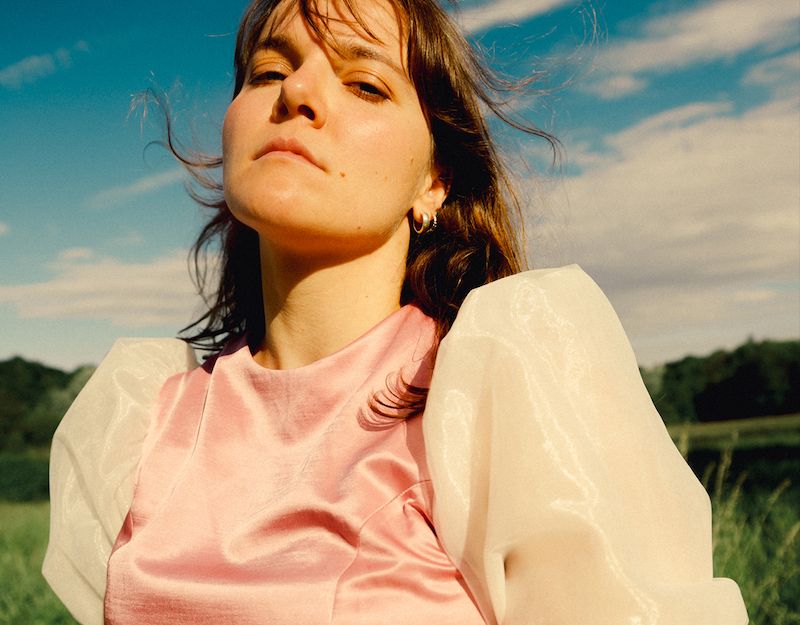 Frau in rosa Kleid mit Puffärmeln steht auf einer Wiese vor blauem Himmel.