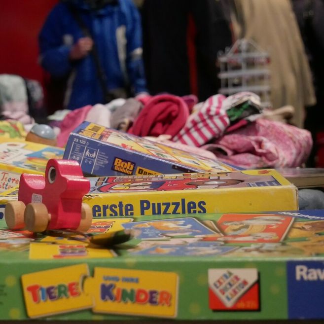Kinderflohmarkt mit Spielzeug, Puzzles und Kleidung; im Vordergrund ein rotes Bobbycar aus Holz, im Hintergrund unscharf stehende Personen.