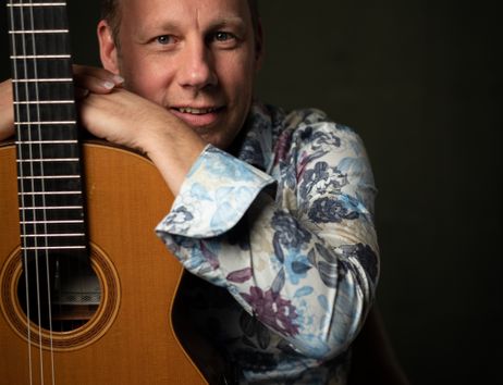 Ein Mann lehnt sich an eine klassische Gitarre und lächelt in die Kamera.