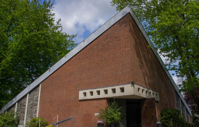 Eine moderne Backsteinkirche mit schrägem Dach und geometrischen Fenstern, umgeben von Bäumen.