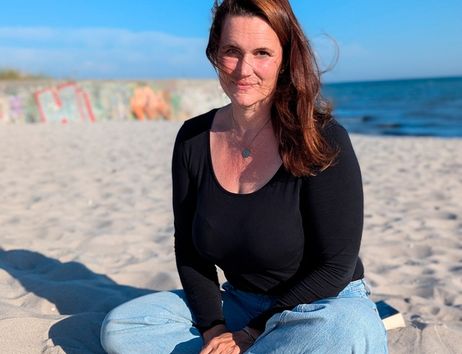 Eine Frau sitzt im Schneidersitz am Strand mit blauem Himmel im Hintergrund.
