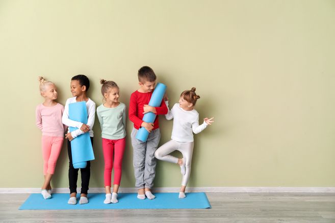 Fünf Kinder stehen auf einer blauen Yogamatte vor einer hellgrünen Wand und halten Yogamatten oder machen eine Balance-Übung.