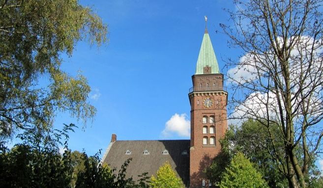 Ein Kirchturm mit grünem Dach ragt in den blauen Himmel, umgeben von Bäumen.