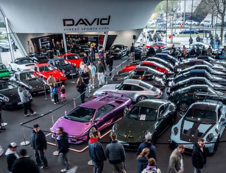 Zahlreiche Sportwagen und Oldtimer, darunter Porsche-Modelle und ein lila Supersportwagen, stehen dicht an dicht vor dem Showroom von DAVID Finest Sports Cars, umgeben von Besucherinnen und Besuchern.