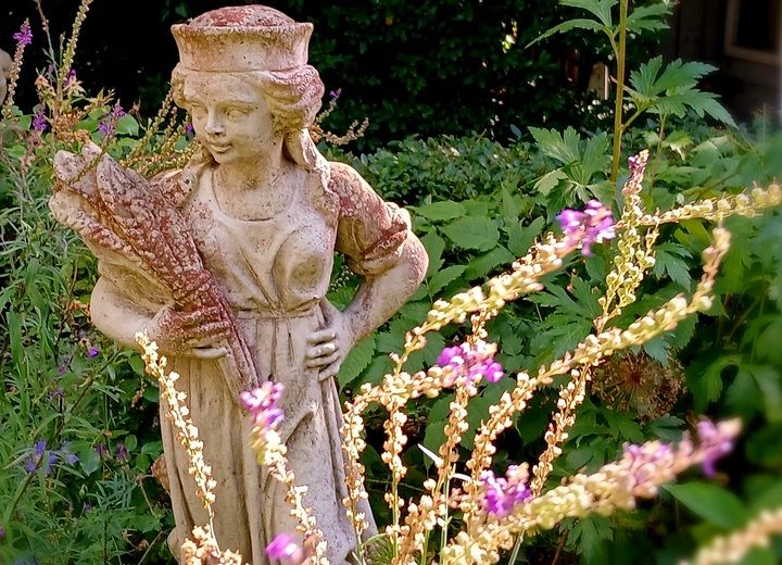 Steinstatue einer Frau mit Blumenstrauß in einem Garten, umgeben von grünen Pflanzen und lila Blüten.