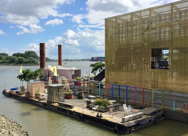 Schwimmende Plattform mit Sitzgelegenheiten und Pflanzen am Fluss, daneben modernes Gebäude mit goldener Fassade.