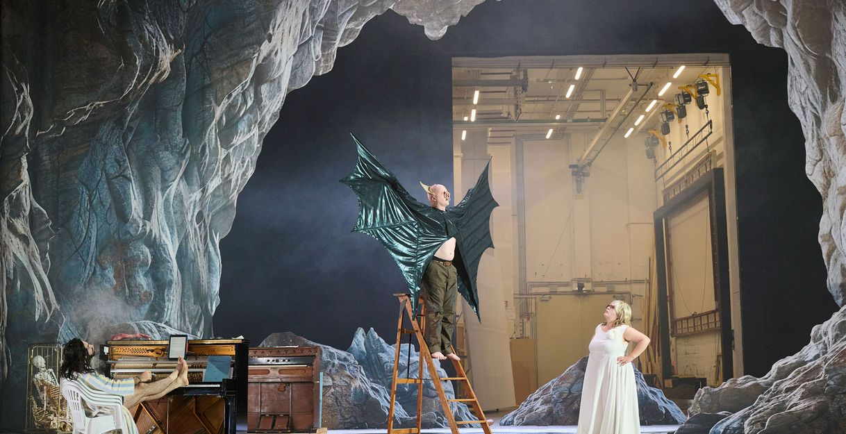 Bühnensituation. Ein Mann auf einer Leiter, die Arme zur Seite ausgestreckt, er trägt ein Kostüm mit großen schwarzen ledrigen Flügeln. Am Boden eine Frau im langen weißen Kleid, sie schaut zu ihm hoch. Links ein Mann am Klavier, die Füße auf die Tasten gelegt. Im Hintergrund eine Höhlenlandschaft. Die offene Hinterbühne ist zu sehen.