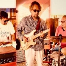 Drei Musiker spielen zusammen: links ein Keyboarder, in der Mitte ein Gitarrist und rechts ein Schlagzeuger.