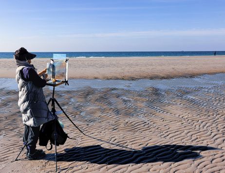 Eine Frau steht mit einer Staffelei am Strand und malt auf eine Leinwand.