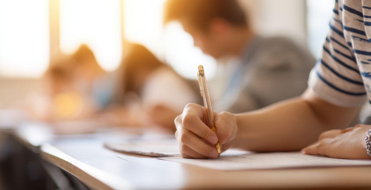 Nahaufnahme einer Person, die mit einem Stift eine Prüfung oder Klassenarbeit schreibt, im unscharfen Hintergrund weitere sitzende Teilnehmende in einem Unterrichtsraum