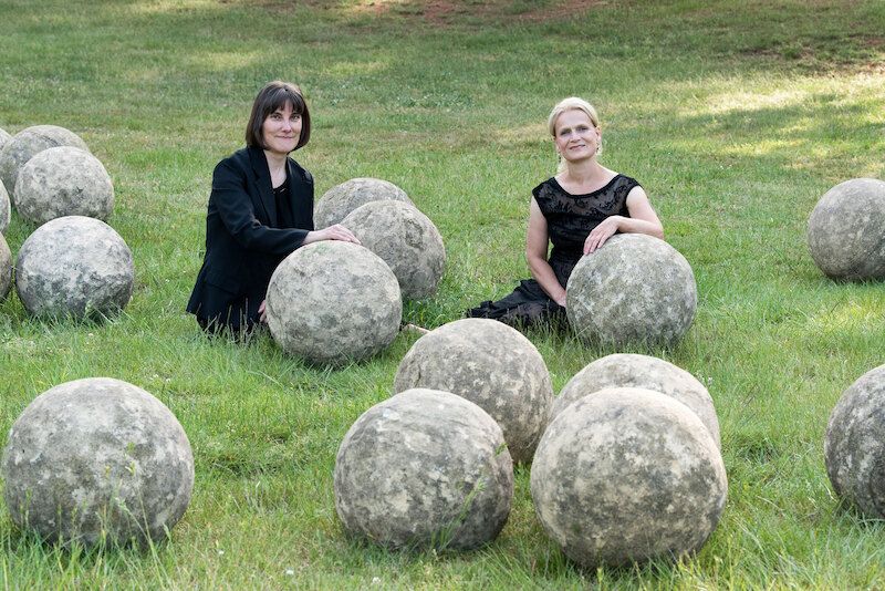 Zwei Frauen sitzen auf einer Wiese zwischen großen Steinkugeln.