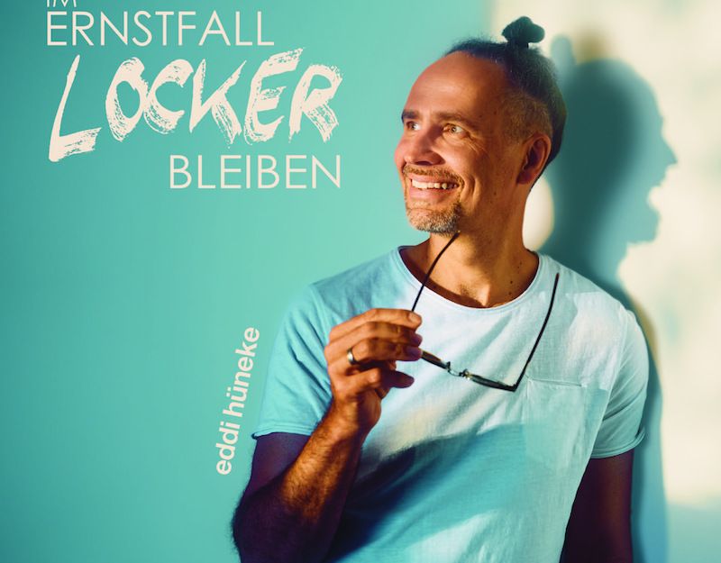 Mann in blauem T-Shirt hält Brille und lächelt vor türkisfarbenem Hintergrund mit Schriftzug 'Im Ernstfall locker bleiben'.