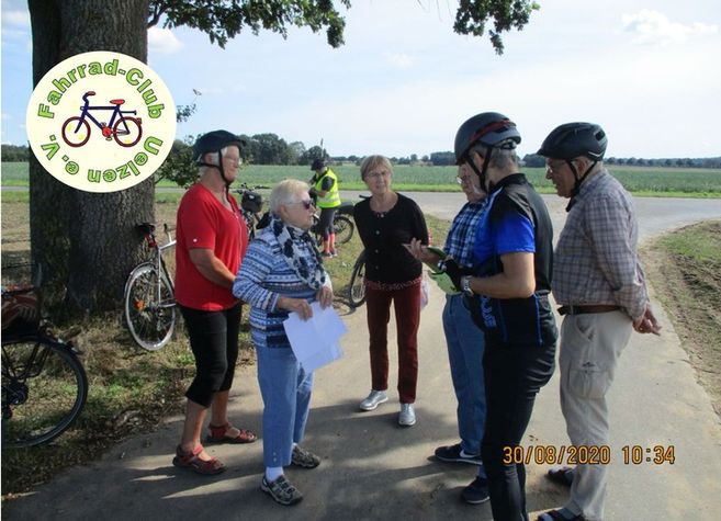 Eine Gruppe von Menschen steht auf einem ländlichen Weg mit Fahrrädern im Hintergrund. Sie tragen Helme und diskutieren miteinander.