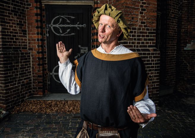 Ein Mann in mittelalterlicher Kleidung steht vor einer Backsteinmauer und einer Holztür.