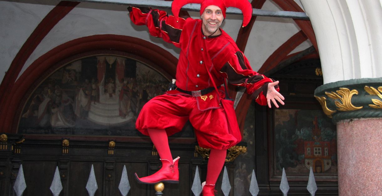 Ein Mann in einem roten Narrenkostüm mit Hörnern posiert auf einem Zaun vor einem historischen Gebäude.
