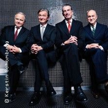 Vier Männer in Anzügen sitzen nebeneinander auf einer Bank vor einem dunklen Hintergrund.