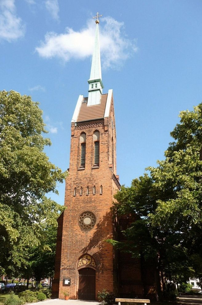 Backsteinkirche mit hohem Turm und grünem Spitzdach, umgeben von Bäumen.