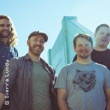 Vier Männer stehen lächelnd vor einem blauen Gebäude unter klarem Himmel.