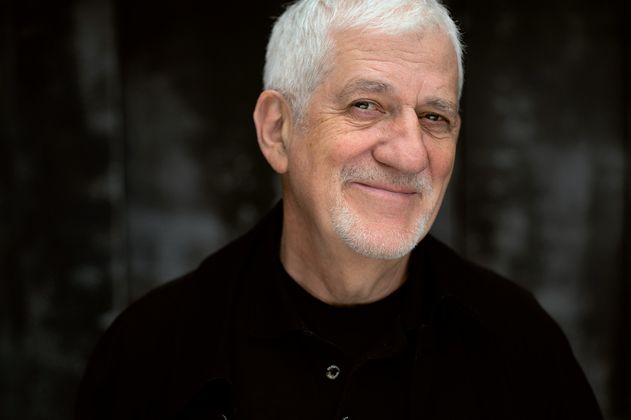 Ein älterer Mann mit weißem Haar und Bart lächelt in die Kamera vor einem dunklen Hintergrund.