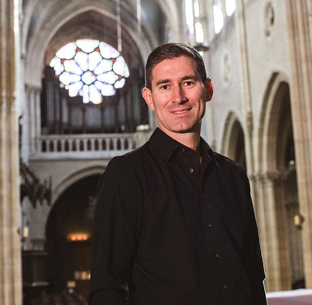 Ein Mann in schwarzem Hemd steht in einer gotischen Kathedrale mit buntem Rosettenfenster im Hintergrund.