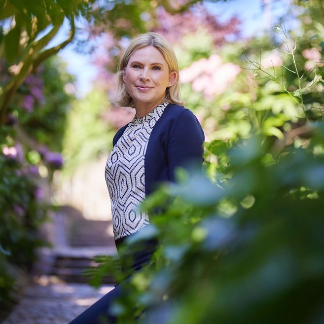 Eine Frau steht in einem sonnigen Garten, umgeben von grünen Blättern und blühenden Pflanzen.