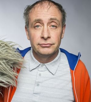 Ein Mann mit grauem Hemd und orange-blauer Jacke schaut direkt in die Kamera. Neben ihm ist ein graues, zotteliges Objekt zu sehen.