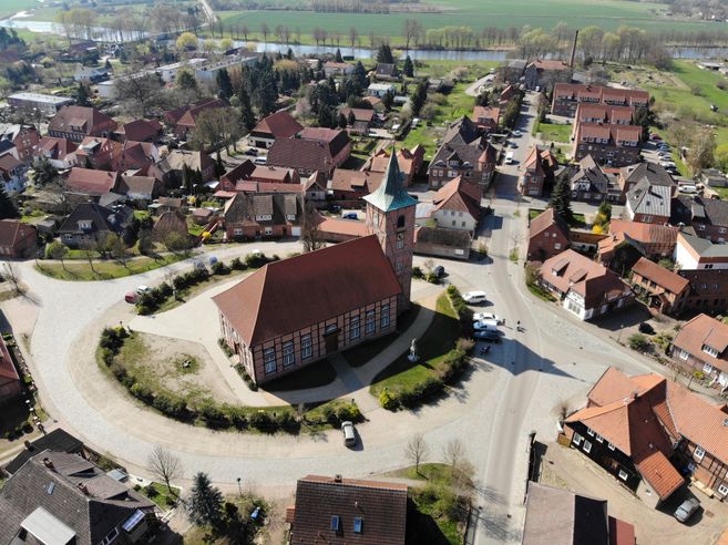Luftaufnahme eines Dorfes mit einer Kirche im Zentrum, umgeben von roten Ziegeldächern und grünen Feldern im Hintergrund.