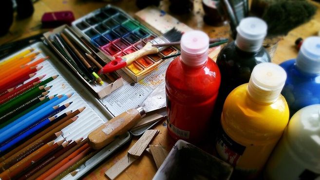 Ein Tisch voller Malutensilien wie Buntstifte, Wasserfarben, Pinsel und Flaschen mit Acrylfarbe in verschiedenen Farben.