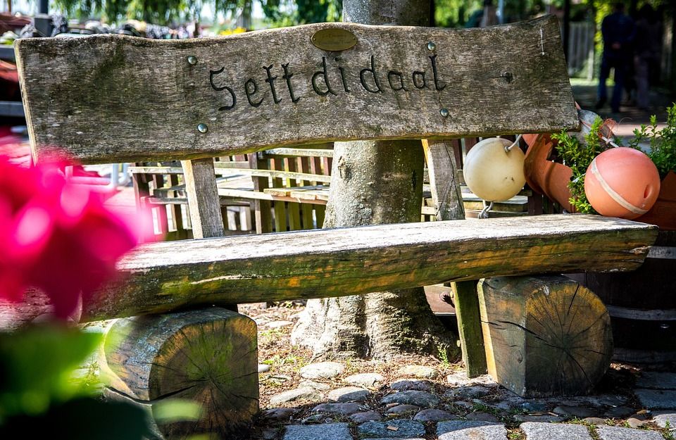 Holzbank mit der Aufschrift 'Sett di doot' im Freien, umgeben von Bäumen und Dekorationen.