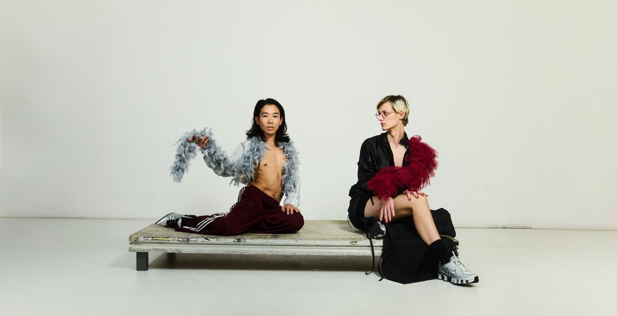 Two performers on a small platform. They wear revealing dark clothing and each wear a feather boa around their shoulders.