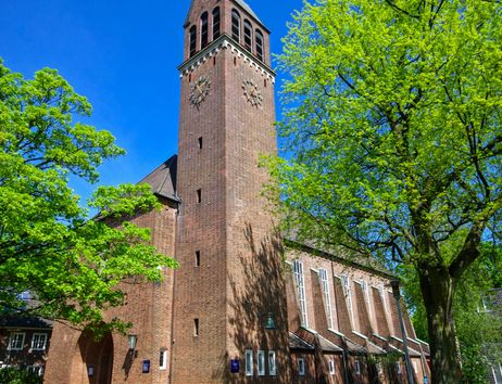 Eine Backsteinkirche mit einem hohen Turm und einer Uhr, umgeben von grünen Bäumen unter einem klaren blauen Himmel.