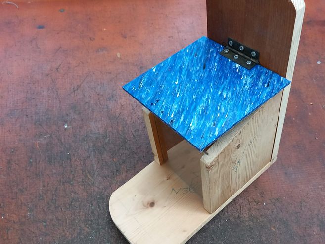 Fertig gebautes Futterhaus aus Holz mit einem blauen Dach aus recycelten Plastik mit Scharnier und einer Scheibe um Nüsse von Außen zu sehen. Das Haus hat davor eine Fläche wo das Eichhörnchen sitzen kann.