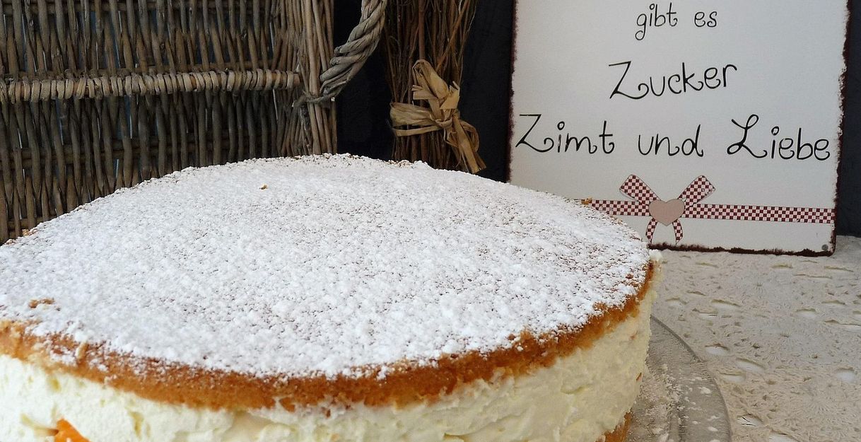 Ein Kuchen mit Puderzucker auf einem Tisch, daneben ein Schild mit der Aufschrift 'In unserer Küche gibt es Zucker, Zimt und Liebe'.