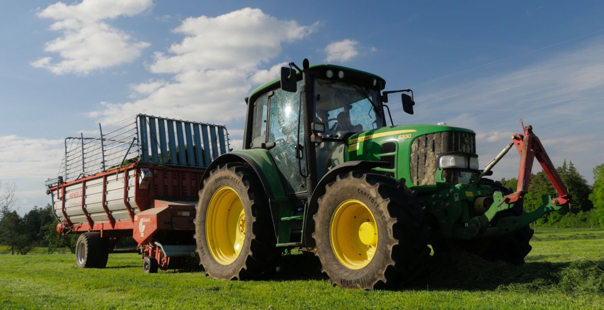 Ein grüner Traktor mit Anhänger steht auf einer Wiese unter blauem Himmel.
